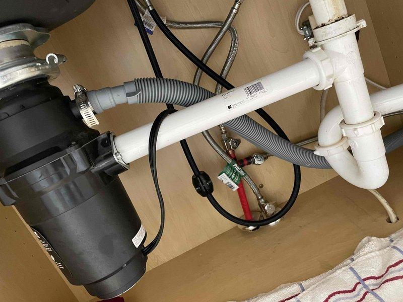 Responded to callback for persistent kitchen sink leak with water now appearing on countertop. Upon inspection, identified leak originating from under-sink plumbing assembly. Examined double-basin stainless steel sink configuration with garbage disposal unit connected to white PVC P-trap drainage system. Tightened slip-joint connections on drain pipes and verified proper sealing at all connection points. Inspected garbage disposal mounting and discharge connections for potential leak sources. Tested water supply lines and checked flexible hose connections for integrity. Ran water through both basins and disposal unit to verify leak repair and confirm no further water intrusion at countertop level or cabinet base. System operating properly upon completion.
