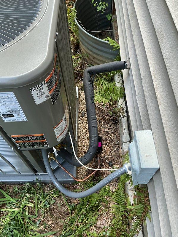 Completed full air conditioning system installation at residential property. Installed new beige condenser unit on gravel pad adjacent to building exterior with proper clearances, ran and insulated refrigerant line set with black foam insulation secured to building, connected electrical disconnect, and performed system evacuation and refrigerant charging using recovery equipment and manifold gauges to manufacturer specifications. All refrigerant lines properly secured and routed to structure, system tested and operational.