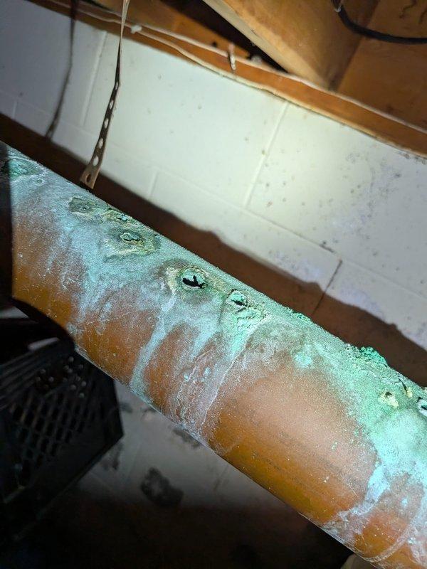 Responded to water leak in basement originating from copper water line connected to water softener. Upon arrival, water was not shut off and leak was actively filling a container at a rate of approximately one gallon per hour. Inspection revealed severely corroded copper piping with extensive green oxidation (verdigris) and white crystalline deposits throughout the affected section. The corrosion had compromised the pipe integrity, resulting in active water seepage. Documented the deteriorated condition of the copper line with photographs showing advanced corrosion around mounting points and along the horizontal run. The pipe exhibited significant environmental wear consistent with age and moisture exposure in the basement environment. Shut off water supply to prevent further water accumulation and mitigate potential water damage to the surrounding area.