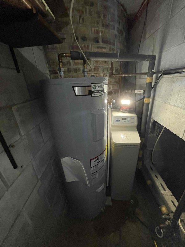 Installed A.O. Smith ENT 60 130 electric storage tank water heater in basement utility room per approved quote. Unit features 4500-watt heating element with 3380-volt capacity, copper piping connections at top, and white plastic tubing for proper drainage. Water heater was positioned adjacent to existing water softener system in narrow utility space with concrete block walls, ensuring adequate clearance for service access and code compliance.