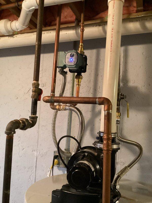 Responded to service call for constant water leak around vent area of 10-year-old tanked water heater. Upon inspection, identified corrosion and rust staining around circulator pump mounting point on top of water heater tank, indicating active leak at pump connection. Documented hydronic heating recirculation system with black circulator pump, copper piping, brass fittings, and flexible stainless steel braided connectors. Photographed evidence of water damage and mineral deposits around pump base where it interfaces with tank surface. Assessed overall plumbing configuration including PVC drainage, copper supply lines, and system controls with digital display indicator.