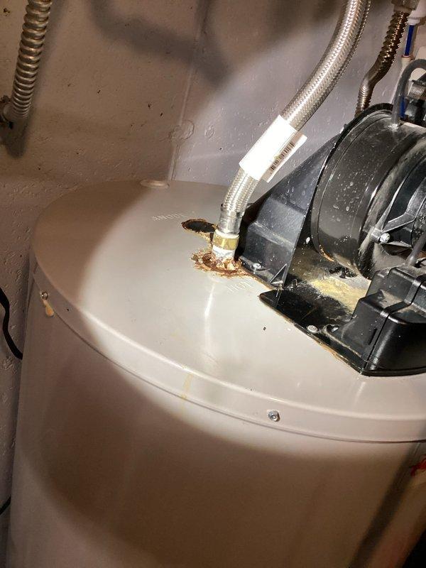 Responded to service call for constant water leak around vent area of 10-year-old tanked water heater. Upon inspection, identified corrosion and rust staining around circulator pump mounting point on top of water heater tank, indicating active leak at pump connection. Documented hydronic heating recirculation system with black circulator pump, copper piping, brass fittings, and flexible stainless steel braided connectors. Photographed evidence of water damage and mineral deposits around pump base where it interfaces with tank surface. Assessed overall plumbing configuration including PVC drainage, copper supply lines, and system controls with digital display indicator.