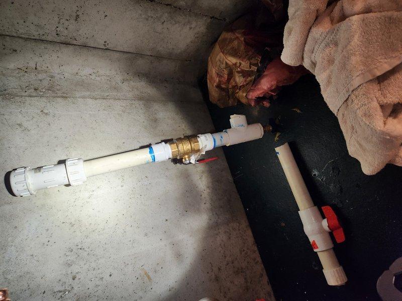 Responded to active water leak in basement with standing water present. Upon arrival, water supply was already shut off by customer. Inspected plumbing system and identified leak source in copper piping connected to sump pump assembly. System includes pump with clear housing, multiple copper lines with red-handled shut-off valves, and white PVC discharge piping with brass fittings and check valve. Documented existing installation showing proper configuration of shut-off valves and pump connections. Assessed standing water conditions and verified water supply remained isolated to prevent further flooding while awaiting repair authorization.