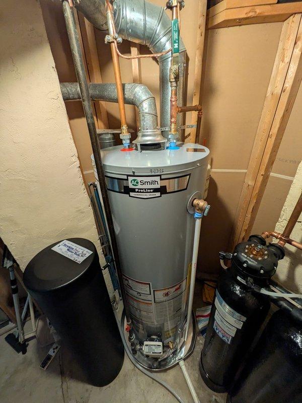 Removed and replaced existing 40-gallon residential water heater with new AO Smith ProLine unit in basement utility area. Installation included connecting metallic ductwork for proper venting, securing copper supply lines with brass fittings, and integrating blue plastic connections at top of unit. Verified proper operation of existing auxiliary equipment including black expansion tank and water treatment system with associated copper piping and valve assemblies.