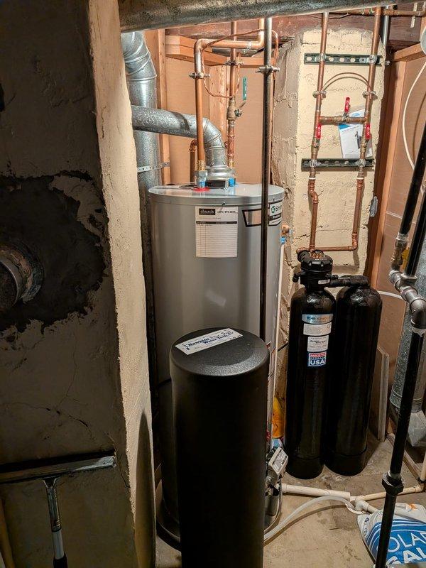 Removed and replaced existing 40-gallon residential water heater with new AO Smith ProLine unit in basement utility area. Installation included connecting metallic ductwork for proper venting, securing copper supply lines with brass fittings, and integrating blue plastic connections at top of unit. Verified proper operation of existing auxiliary equipment including black expansion tank and water treatment system with associated copper piping and valve assemblies.