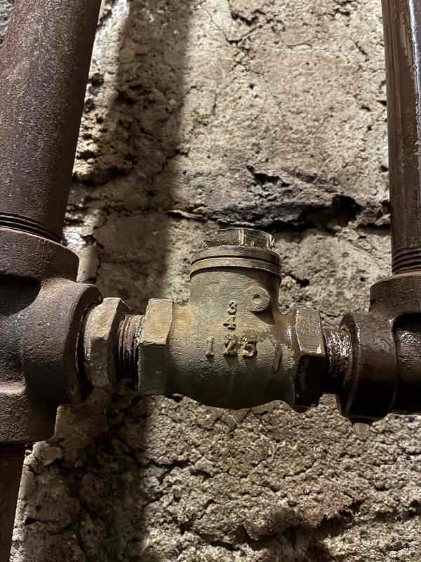 Responded to emergency no-heat call for a radiant heating system with a boiler less than 5 years old. Upon inspection, discovered a major leak in an unidentified line (either supply or return) located in the garage. Documented the condition with photos of the plumbing connections and valve assembly for further assessment.