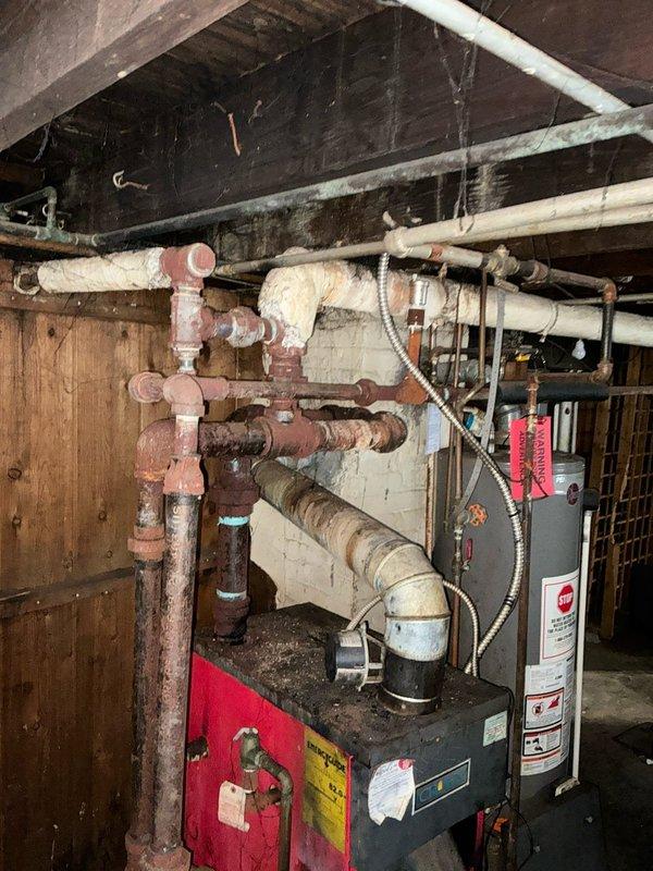 Conducted comprehensive assessment of aging boiler system for replacement estimate. Observed significant deterioration including extensive corrosion on iron distribution pipes, compromised pipe insulation, and multiple connection points showing advanced oxidation. System appears to be non-operational with visible red tag indicating potential safety concerns or previous service notification.