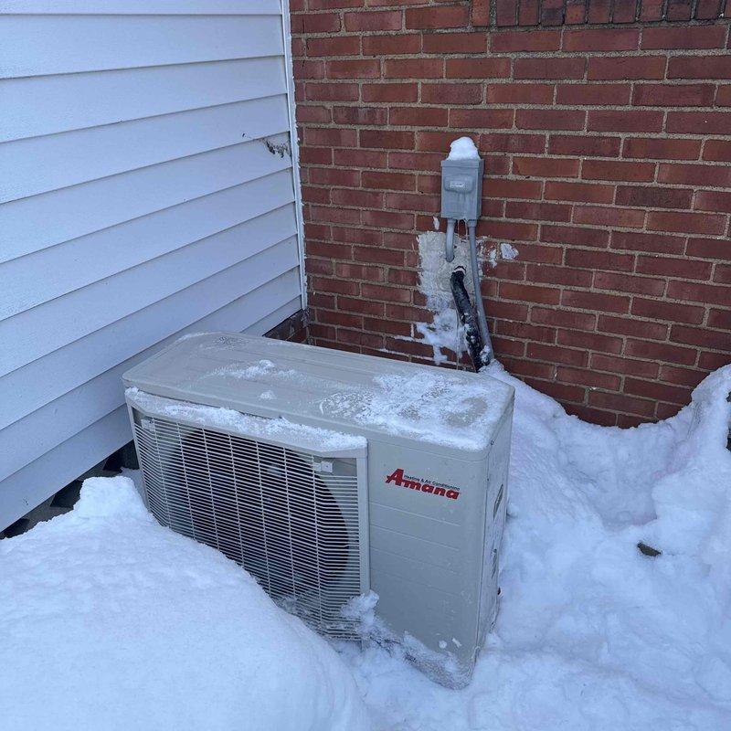 Performed warranty service call to install correct thermostat with Palmer and inspect outdoor Amana air conditioning unit. Noted significant snow accumulation around and on top of the unit, with snow partially burying the equipment and some ice buildup on the conduit and wall near the electrical disconnect box.