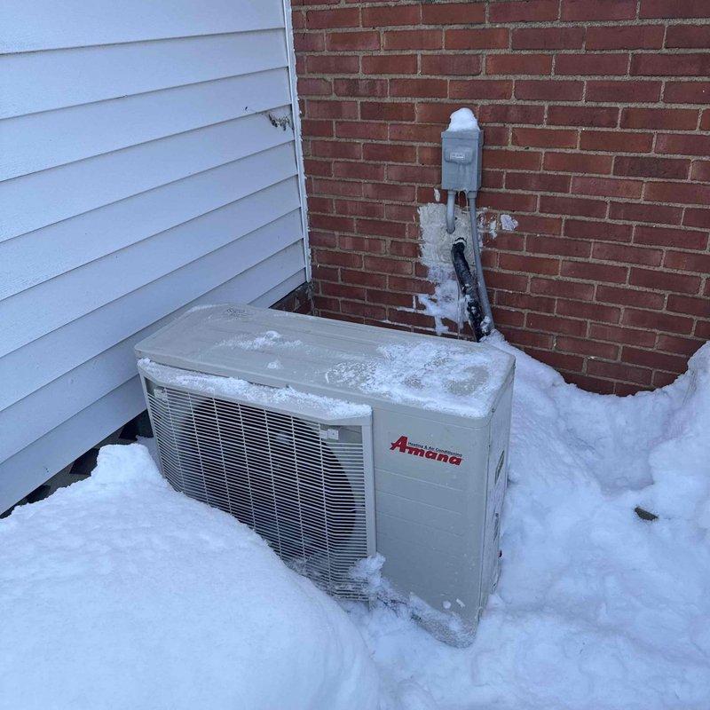 Performed warranty service call to install correct thermostat with Palmer and inspect outdoor Amana air conditioning unit. Noted significant snow accumulation around and on top of the unit, with snow partially burying the equipment and some ice buildup on the conduit and wall near the electrical disconnect box.