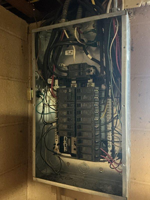 Performed electrical inspection of service panel in basement area. Inspected circuit breaker panel mounted on concrete block wall, examining wiring connections and breaker functionality. Also observed water pump/valve assembly with PVC piping connections adjacent to utility tank.