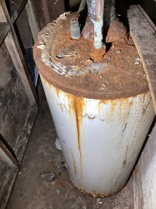 Conducted inspection of water heater system exhibiting severe corrosion. Unit shows extensive rust formation throughout the tank body with significant deterioration at connection points and top surface. Observed water/moisture intrusion at pipe connections and rust streaks extending down tank sides, indicating ongoing water issues accelerating the corrosion process.