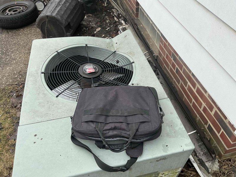 Inspected outdoor AC condenser unit damaged by fallen gutter. Unit shows weathering and impact damage but appears functionally intact. Documented condition of surrounding area including brick foundation wall and electrical connections to assess replacement options.