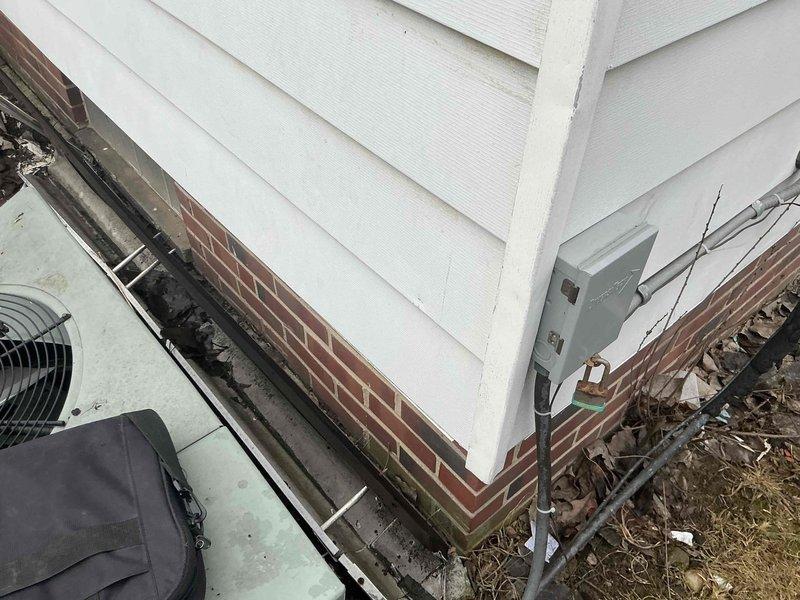 Inspected outdoor AC condenser unit damaged by fallen gutter. Unit shows weathering and impact damage but appears functionally intact. Documented condition of surrounding area including brick foundation wall and electrical connections to assess replacement options.