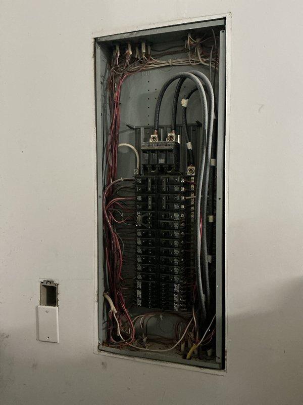 Conducted comprehensive electrical inspection, photographing and documenting an exposed wall outlet requiring installation completion and examining the circuit breaker panel which showed organized but crowded wiring with multiple red/white conductors and black cables entering from the top.