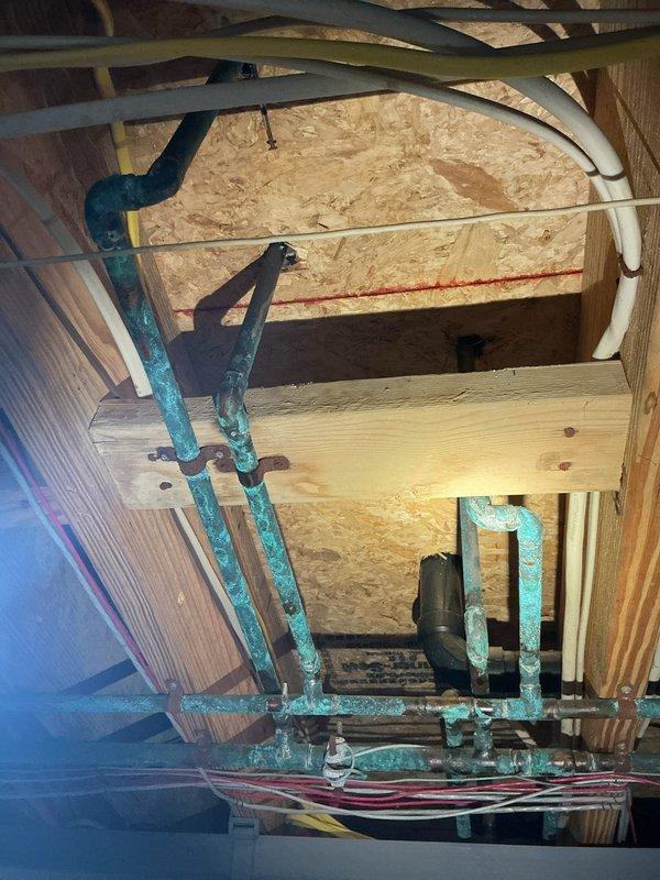 Conducted comprehensive plumbing inspection focusing on aging copper supply lines exhibiting significant verdigris and corrosion throughout the unfinished space. Documented condition of plumbing infrastructure including copper pipes mounted to wooden structural elements, adjacent PVC lines, and associated mounting hardware to assess system integrity and identify potential failure points.