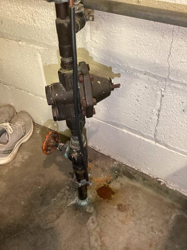 Conducted plumbing inspection of water supply system in utility area. Identified significant corrosion at pipe-floor junction with visible greenish-blue oxidation and orange staining on concrete, indicating active leak or condensation issues. Documented condition of pressure regulator/valve assembly and electrical conduit installation for follow-up service.
