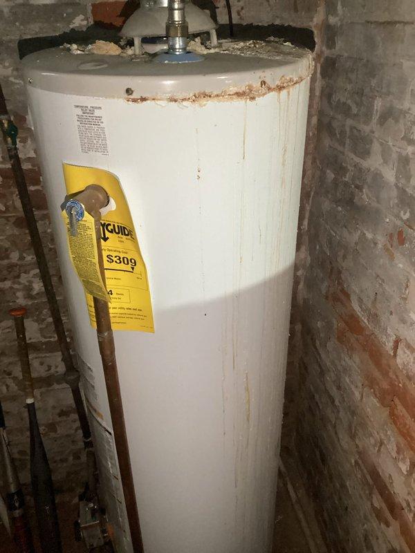 Completed comprehensive plumbing inspection of residential water heater system and documented current condition with photographic evidence. Water heater exhibited visible signs of aging including rust corrosion around upper rim and pressure relief valve assembly, peeling paint on surrounding brick wall, and sediment accumulation at base, with all supply lines, copper piping, reflective insulation wrapping, and flexible braided connections visually assessed for immediate safety concerns.