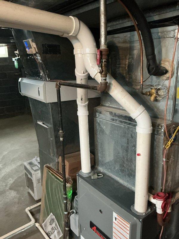 Responded to customer report of furnace water leak in basement mechanical room. Upon inspection, examined gray metal furnace unit with white PVC condensate drainage piping system running vertically and horizontally with multiple elbow joints. Inspected all condensate drain connections, pipe penetrations through wall surfaces, and drain line routing. Photographed installation showing PVC drain line secured with metal straps at wall penetration point, noting patching material and sealing compound around pipe opening. Verified proper drainage slope and checked for blockages in condensate system. Documented equipment layout including furnace, water heater, and dehumidifier unit to assess potential leak sources and drainage pathways.