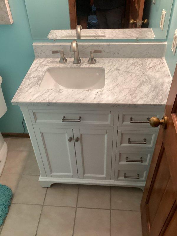 Completed installation of customer-supplied bathroom vanity unit featuring white cabinetry with marble-patterned countertop and undermount sink. Connected brushed nickel faucet fixture with dual-handle configuration and installed matching marble backsplash. Performed sump pump maintenance in utility area. Removed and disposed of existing pressure reducing valve (PRV) and water softener system, including disconnection of copper supply lines and proper capping of plumbing connections.