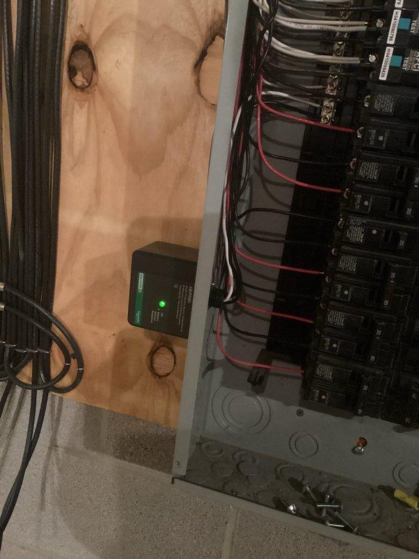 Installed whole home surge protection device at main electrical panel. Device mounted on plywood wall adjacent to 200-amp main breaker panel, with proper wiring connections made to protect entire electrical system from voltage spikes and power surges. Unit operational with green indicator light confirming active protection status.
