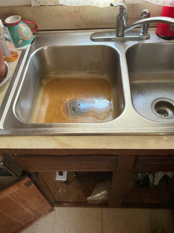 Performed comprehensive plumbing inspection of kitchen sink and drainage system. Diagnosed completely clogged garbage disposal unit and standing water issue in left basin. Documented under-sink plumbing configuration including disposal unit, PVC trap assembly, and cabinet condition for recommended repairs.