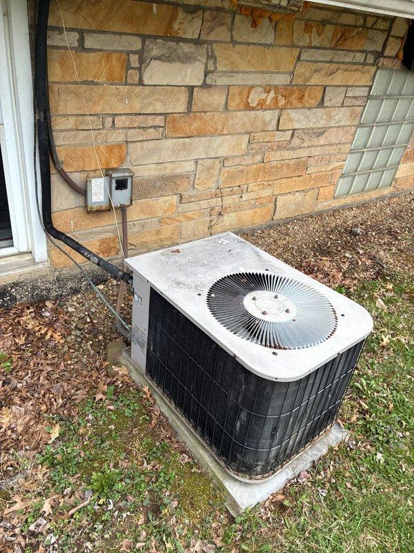 Performed site evaluation and equipment assessment for air conditioning system replacement. Existing 15-year-old Goodman unit evaluated for replacement with Trane equipment per customer request. Documented current installation including outdoor condensing unit positioned on concrete pad against stone veneer exterior wall, electrical disconnect, and conduit routing. Photographed equipment location, surrounding area conditions, and exterior building features to support replacement proposal specifications.