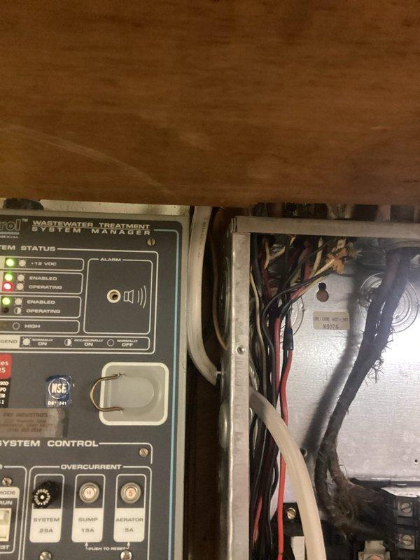 Installed and wired wastewater treatment system control panel in utility area, including mounting control manager unit with LED status indicators and system control functions beneath floor joists. Completed electrical connections in adjacent junction box with proper wire routing and overcurrent protection circuits rated for 25A, 15A, and 8A loads. Routed exterior electrical wiring along soffit to support system operation.
