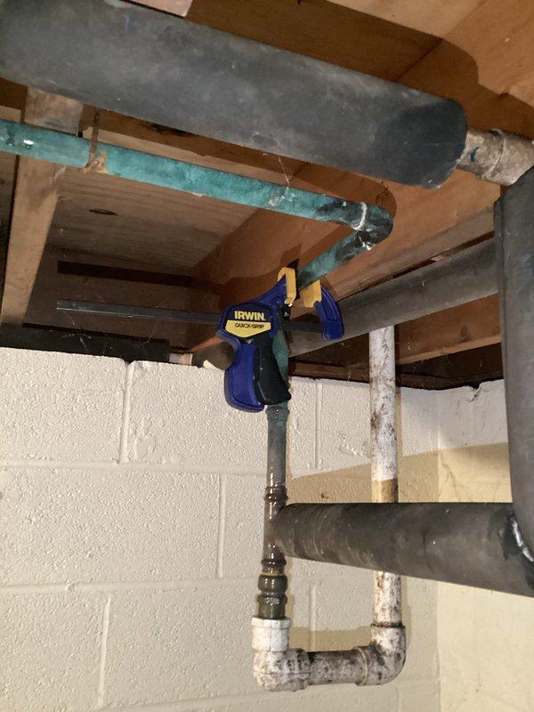 Responded to service call for leaking pipe in geothermal system. Upon inspection, located compromised piping in basement utility area where geothermal loop connects to system infrastructure. Documented existing conditions showing turquoise PVC piping with visible age-related deterioration and adjacent gray ductwork components. Temporarily stabilized affected section using bar clamp to secure piping and prevent further water loss until permanent repairs can be scheduled. Customer advised that replacement of deteriorated pipe section will be required to restore system integrity.