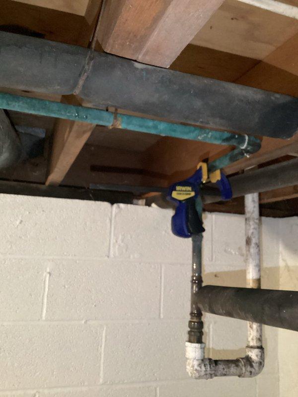 Responded to service call for leaking pipe in geothermal system. Upon inspection, located compromised piping in basement utility area where geothermal loop connects to system infrastructure. Documented existing conditions showing turquoise PVC piping with visible age-related deterioration and adjacent gray ductwork components. Temporarily stabilized affected section using bar clamp to secure piping and prevent further water loss until permanent repairs can be scheduled. Customer advised that replacement of deteriorated pipe section will be required to restore system integrity.