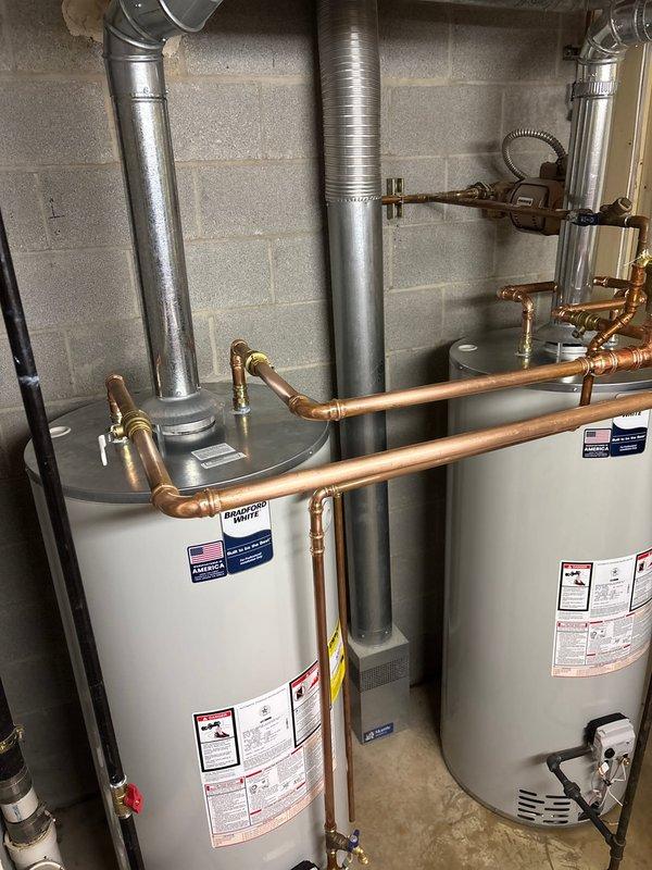 Completed installation of twin 75-gallon atmospheric Bradford White water heaters in residential utility space. Units were positioned side by side against concrete block wall with professional copper piping connections, brass fittings, and shut-off valves installed throughout the system. Metallic ductwork and ventilation components were properly integrated, including flexible aluminum ducting and rigid vent piping with elbow joints. All plumbing connections were secured with appropriate fittings, and the installation was tested and verified for proper operation.