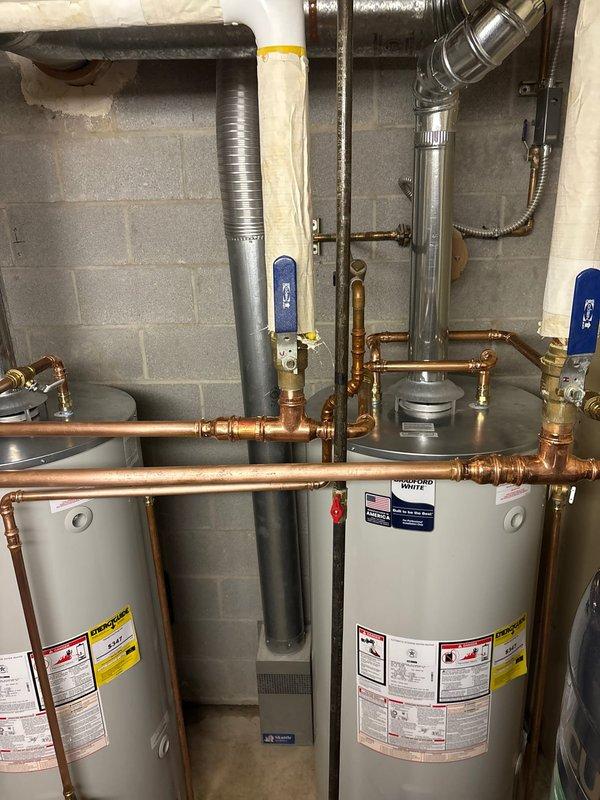 Completed installation of twin 75-gallon atmospheric Bradford White water heaters in residential utility space. Units were positioned side by side against concrete block wall with professional copper piping connections, brass fittings, and shut-off valves installed throughout the system. Metallic ductwork and ventilation components were properly integrated, including flexible aluminum ducting and rigid vent piping with elbow joints. All plumbing connections were secured with appropriate fittings, and the installation was tested and verified for proper operation.