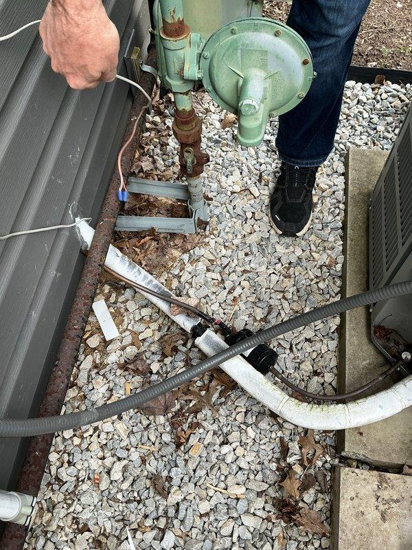 Responded to service call for air conditioning system following condenser relocation due to siding installation. Customer reported having pumped refrigerant into condenser unit prior to moving equipment. Upon arrival, performed visual inspection of relocated condenser unit and associated refrigerant lines. Observed condenser positioned on gravel pad with turquoise-green compressor housing and connecting piping showing weathering and age. Noted presence of refrigerant lines including white insulated suction line and corrugated flexible hose routing. Assessed condition of copper line connections and isolation valves on unit. Documented equipment positioning and line routing with photographs for service records. System requires proper refrigerant recovery, line set evaluation, and reinstallation procedures to restore operation safely per EPA regulations and manufacturer specifications.