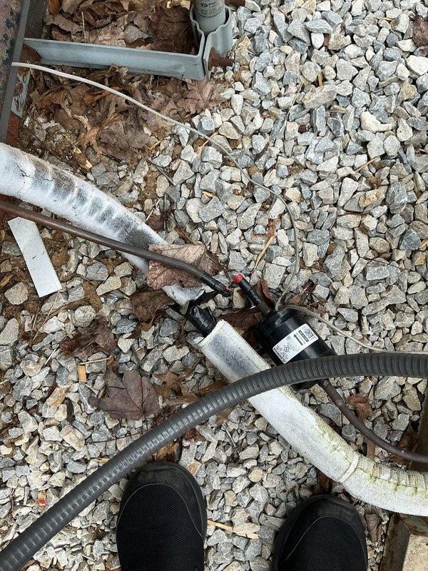 Responded to service call for air conditioning system following condenser relocation due to siding installation. Customer reported having pumped refrigerant into condenser unit prior to moving equipment. Upon arrival, performed visual inspection of relocated condenser unit and associated refrigerant lines. Observed condenser positioned on gravel pad with turquoise-green compressor housing and connecting piping showing weathering and age. Noted presence of refrigerant lines including white insulated suction line and corrugated flexible hose routing. Assessed condition of copper line connections and isolation valves on unit. Documented equipment positioning and line routing with photographs for service records. System requires proper refrigerant recovery, line set evaluation, and reinstallation procedures to restore operation safely per EPA regulations and manufacturer specifications.