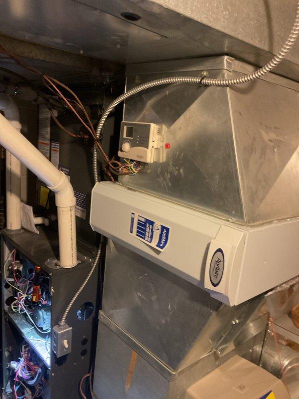 # HVAC Heating System Maintenance Completed

Performed comprehensive heating maintenance on residential HVAC system located in mechanical room. Conducted thorough inspection of furnace cabinet, blower motor assembly, and associated electrical components. Verified proper operation of humidifier unit with transparent cover, confirming water supply and drainage functionality. 

Inspected all electrical connections including terminal blocks with blue and orange wiring on air handler cabinet. Examined blower motor assembly featuring black cylindrical component with ribbed sections and verified secure connections of multi-colored wiring harnesses throughout system. Checked green motor unit and associated capacitors mounted on equipment panel for proper operation.

Verified integrity of PVC condensate drain lines and copper refrigerant lines for proper routing and connections. Inspected flexible metal conduits protecting electrical wiring runs. Examined control panel and thermostat mounted on