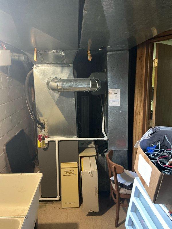 Conducted on-site assessment of existing HVAC system in basement utility area to provide replacement options. Documented current Lennox furnace installation with metal flue pipe and associated ductwork configuration, including black air handler component and horizontal cylindrical duct connections. Photographed equipment layout showing furnace positioned against concrete block wall with Rheem water heater adjacent, noting PVC plumbing lines and standard gas line connections with proper venting systems in place.
