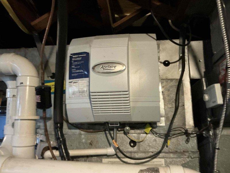 Documented existing HVAC system configuration in basement mechanical room, including Aprilaire humidifier unit with associated PVC plumbing and electrical connections. Photographed current equipment installation showing white PVC piping with metallic tape repair section, concrete block wall mounting, and ductwork connections to provide comprehensive visual reference for replacement system specifications and equipment upgrade options.