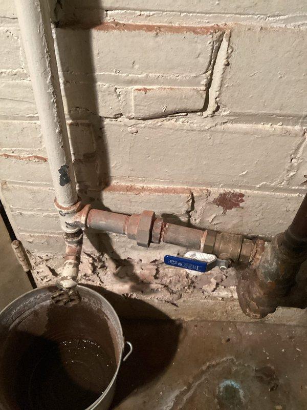 Responded to customer complaint of low water pressure affecting multiple faucets throughout the home. Upon investigation, discovered significant corrosion and mineral buildup in the main water supply lines located in the basement utility area. Identified rust-colored staining and oxidation on metal pipes and fittings, indicating advanced deterioration of the plumbing system. Documented corroded piping with visible scale accumulation and discolored water present in collection bucket, confirming sediment contamination in the water supply. Assessed blue-handled ball valve and surrounding pipe connections showing signs of aging and potential flow restriction. Exterior inspection revealed moisture intrusion at foundation level with algae growth on brick surfaces, suggesting possible water supply line issues contributing to pressure problems. Photographed affected areas for documentation and recommended comprehensive evaluation of water supply system to address corrosion-related flow restric