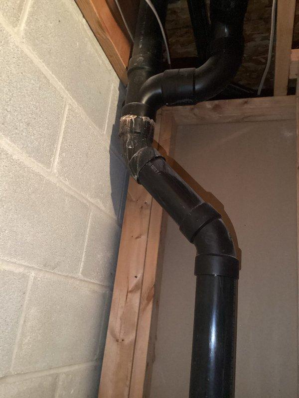 Conducted on-site assessment for complete sump pump failure with unit showing no operational function. Documented existing drainage system configuration including black PVC discharge piping running through basement framing with multiple elbows and connections secured with metal hose clamps. Photographed current installation showing drainage lines integrated with structural wooden framing and foundation walls. Provided comprehensive estimate for full sump pump replacement to restore proper basement water removal capability.