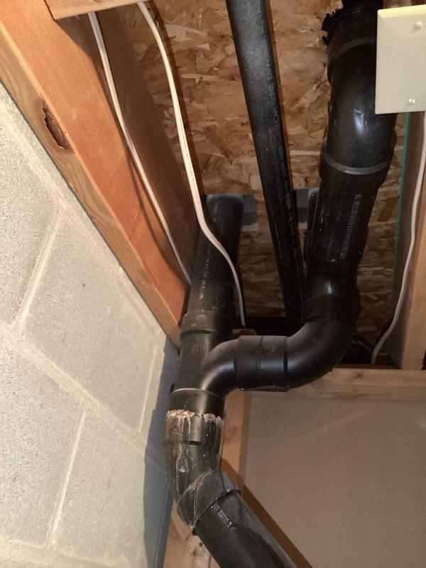 Conducted on-site assessment for complete sump pump failure with unit showing no operational function. Documented existing drainage system configuration including black PVC discharge piping running through basement framing with multiple elbows and connections secured with metal hose clamps. Photographed current installation showing drainage lines integrated with structural wooden framing and foundation walls. Provided comprehensive estimate for full sump pump replacement to restore proper basement water removal capability.