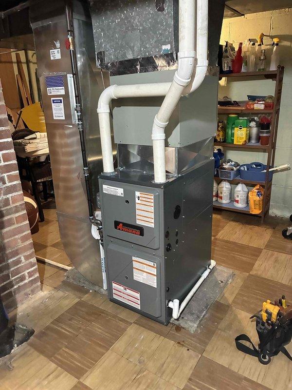 Completed installation of new Amana 96% AFUE high-efficiency gas furnace in basement utility area. System includes PVC venting routed through existing ductwork connections, gas piping with black iron threaded fittings, and unit mounted on concrete pad with proper clearances maintained. All connections tested and operational, with furnace commissioned and verified for proper heating cycle operation.