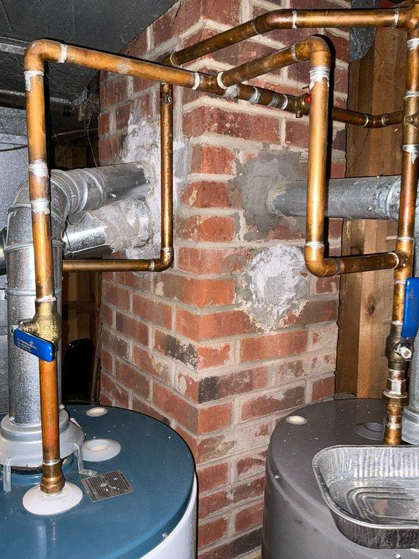 Conducted on-site evaluation for tankless water heater installation in residential property. Assessed existing water heating system consisting of traditional tank-style water heater located in basement utility area with copper supply lines routed around brick chimney structure. Documented current plumbing configuration including copper piping layout, shut-off valve locations on exterior, and available space for potential tankless unit installation. Photographed existing equipment and plumbing infrastructure to develop accurate installation proposal and determine necessary modifications to gas lines, venting, and water supply connections for tankless conversion.