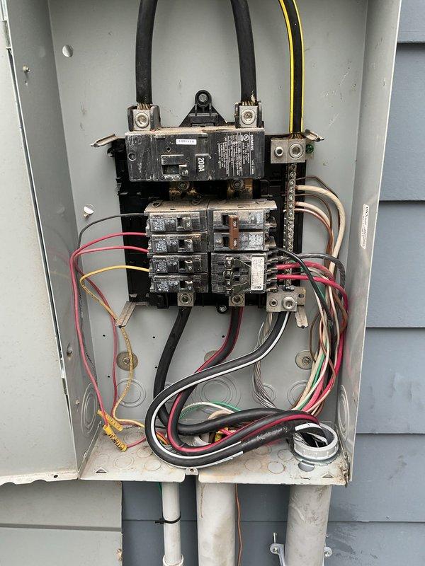 # Partial Rewire - 200 Amp Service Upgrade

Completed a comprehensive partial rewire with 200 amp service upgrade involving replacement and installation of two electrical panels. The interior panel was surface-mounted on wooden backing in the basement utility area, featuring dual circuit breaker arrays with proper color-coded wiring (red, black, white) connected to main bus bars. Three large main power feed cables were terminated at the top of the panel with appropriate connectors.

The exterior panel was mounted on the building's vinyl siding and included a main disconnect switch at the top with dual feed cables entering through the enclosure. Below the disconnect, installed a full complement of circuit breakers arranged in standard residential configuration. Multiple branch circuits were connected using properly sized conductors in various colors including red, black, white, yellow, and green wiring. Gray PVC conduit entries were installed at the bottom of the exterior panel for prot