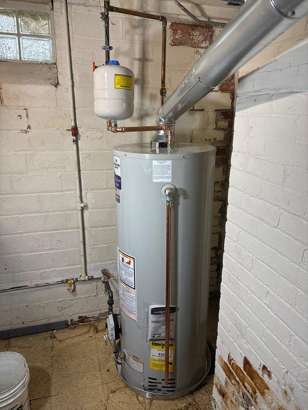 Completed installation of a 50-gallon Bradford White tanked water heater in basement utility area. Unit was properly positioned on tiled floor against cinder block wall foundation, with all copper supply lines connected using brass fittings and shutoff valves. White expansion tank was installed and connected to the cold water inlet with appropriate pressure relief configuration, featuring blue pressure valve at top. All gas connections, venting, and burner assembly were installed per manufacturer specifications, with proper clearances maintained for safe operation and future maintenance access.
