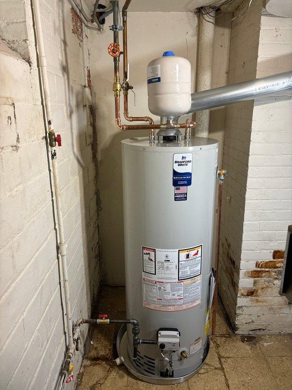 Completed installation of a 50-gallon Bradford White tanked water heater in basement utility area. Unit was properly positioned on tiled floor against cinder block wall foundation, with all copper supply lines connected using brass fittings and shutoff valves. White expansion tank was installed and connected to the cold water inlet with appropriate pressure relief configuration, featuring blue pressure valve at top. All gas connections, venting, and burner assembly were installed per manufacturer specifications, with proper clearances maintained for safe operation and future maintenance access.