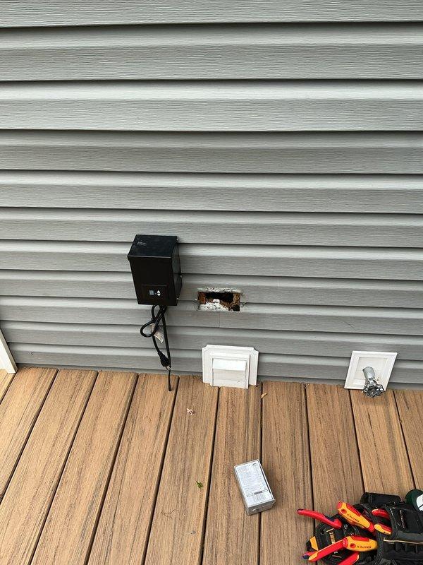 Completed electrical estimate for exterior outlet installation. Assessed existing electrical panel with dual rows of circuit breakers and evaluated wire routing from interior panel through wall to proposed exterior outlet location. Identified penetration point in exterior siding where new weatherproof outlet box will be mounted and documented current conditions including exposed opening and existing utility access panels on deck area.