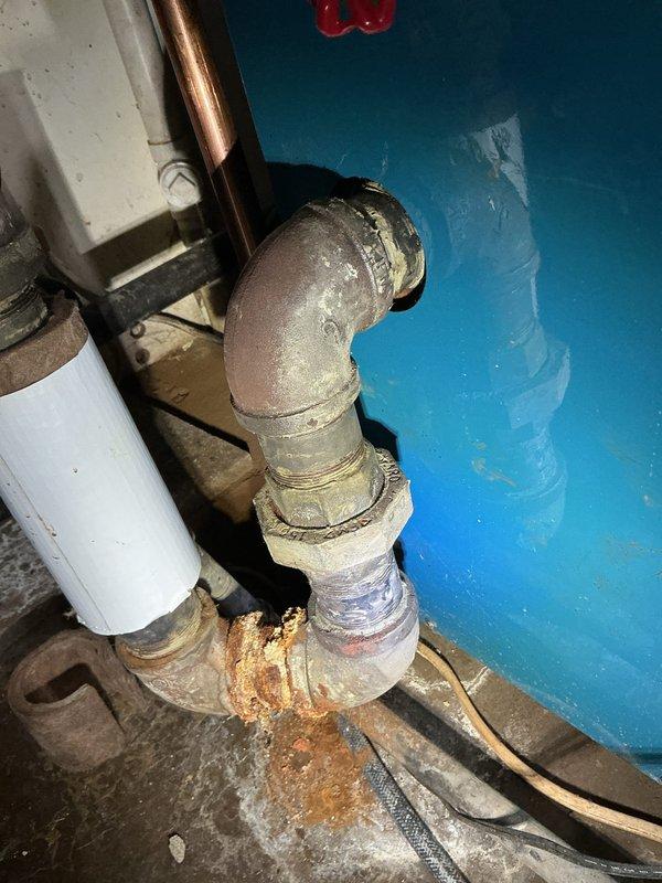 Responded to service call for reported boiler pipe leak. Upon inspection, identified significant corrosion and rust buildup at threaded pipe joint connection on boiler supply piping. Documented existing conditions showing deteriorated gray pipe elbow fitting with orange-brown rust concentration at joint interface and aging plumbing infrastructure with mixed piping materials including PVC, copper, and metal components.
