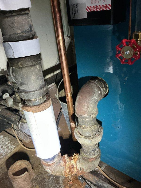 Responded to service call for reported boiler pipe leak. Upon inspection, identified significant corrosion and rust buildup at threaded pipe joint connection on boiler supply piping. Documented existing conditions showing deteriorated gray pipe elbow fitting with orange-brown rust concentration at joint interface and aging plumbing infrastructure with mixed piping materials including PVC, copper, and metal components.