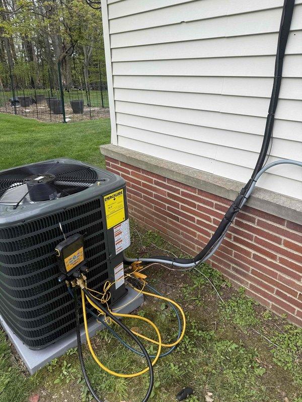 Completed installation of new Goodman air conditioning system on existing concrete pad adjacent to residential structure. Unit positioned on level concrete base with proper clearances from brick foundation and vinyl siding exterior. Performed electrical connections, refrigerant line installation, and system startup procedures. Conducted diagnostic testing using electronic testing equipment to verify proper amperage draw, voltage levels, and refrigerant pressures. System commissioned and operating within manufacturer specifications.