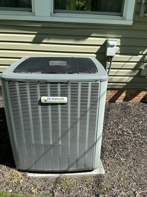 Completed routine cooling system maintenance on residential outdoor condensing unit. Performed comprehensive inspection of P.K. Wadsworth unit including examination of cabinet condition, ventilation louvers, electrical disconnect, and refrigerant lines. Unit found to be in normal operating condition with no visible damage, corrosion, or deterioration present. All components clean and properly maintained with adequate clearance from surrounding landscape materials.