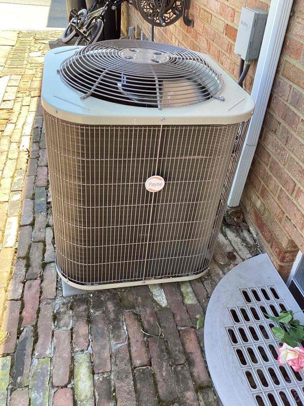 Performed comprehensive cooling season maintenance on residential Payne brand air conditioning system. Inspected condensing unit exterior components including fan assembly, protective grille, condenser coil housing with vertical fins, and electrical connections at wall-mounted junction box. Verified proper unit clearances, stable mounting on brick paver foundation, and refrigerant line connections along exterior wall, confirming system operational readiness for cooling season.